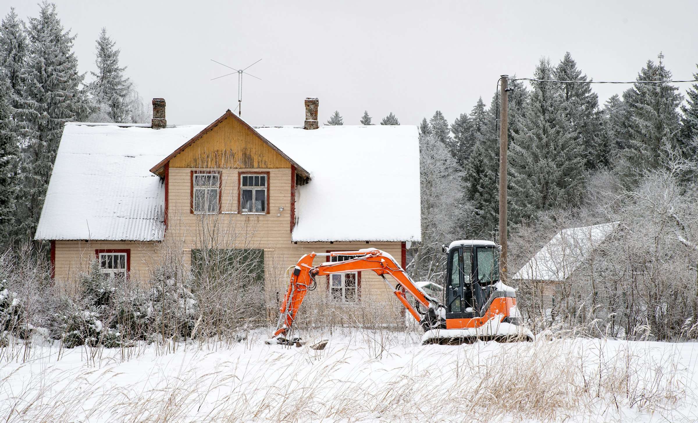 Oranssi kaivinkone lumisella pihalla vanhan puutalon edessä.